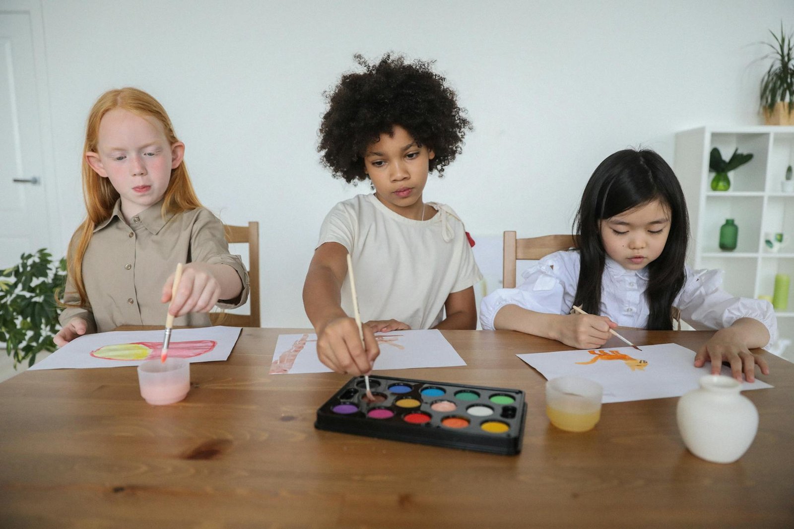 talented diverse little girls painting on papers with watercolors while sitting together at table